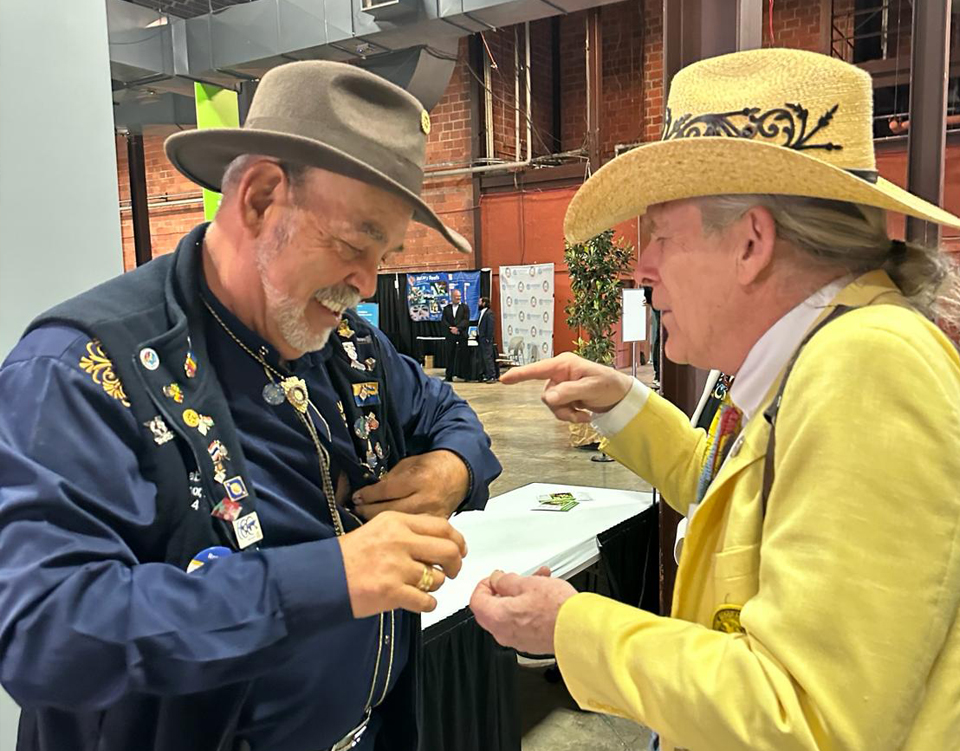 Two men in hats laughing together, one wearing a vest covered in pins.