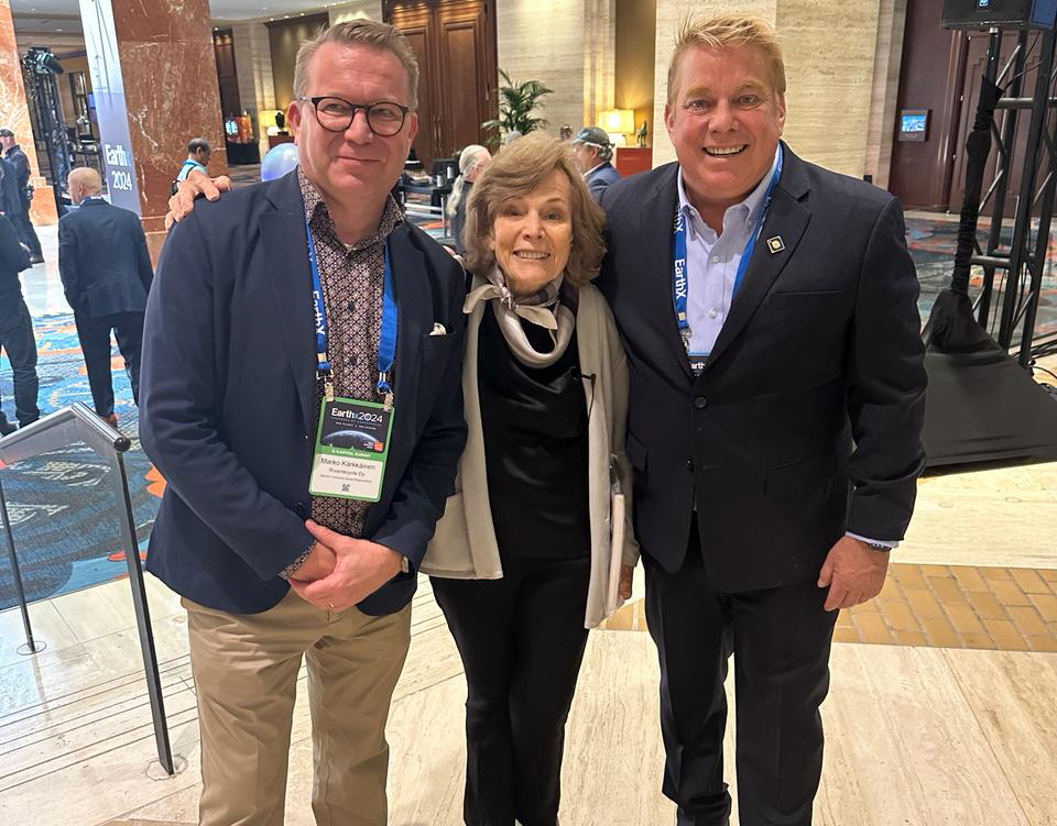 Three attendees with lanyards smiling together in a busy convention hall.