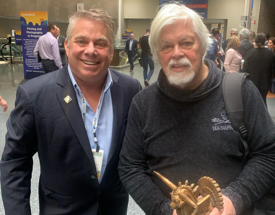 Two men smiling side by side at a convention, one holding a small sculpture.