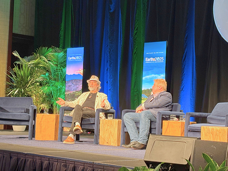 Two speakers seated on a stage with blue lighting and plants, in conversation during a session.