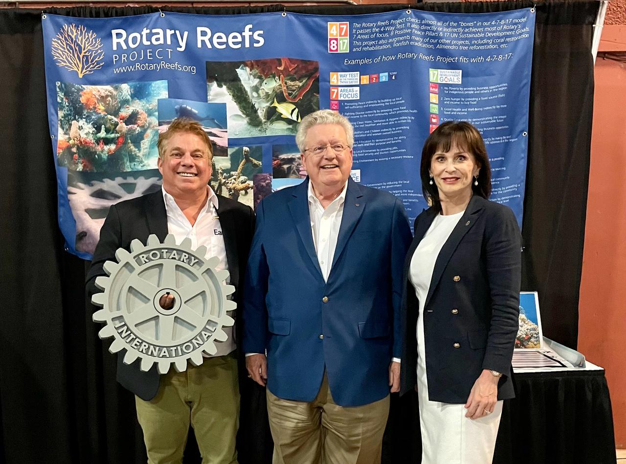 Three Rotary members standing in front of a Rotary Reefs display booth, one holding a Rotary wheel emblem.